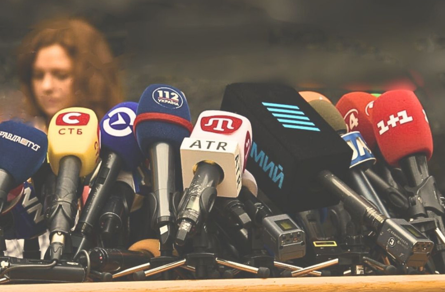 Many microphones on a podium with a woman in the far left background.