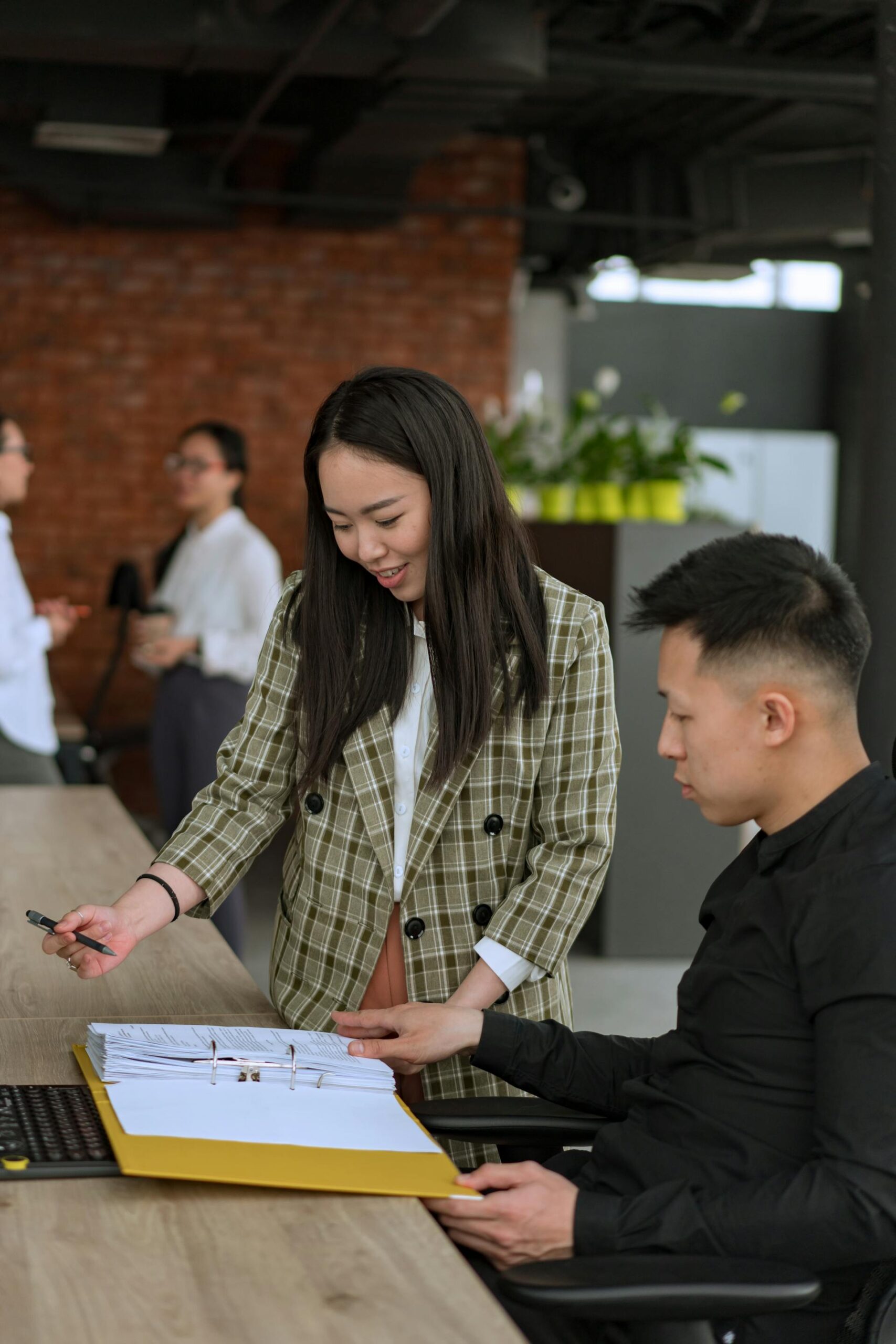 Woman in plaid sportcoat standing and guiding a colleague who is sitting at a long table with a notebook open.